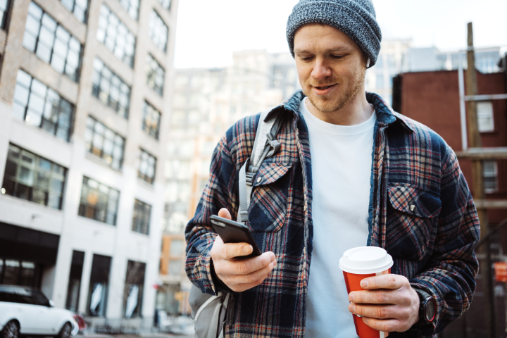man in blue hat and flannel holds coffee and looks at his phone